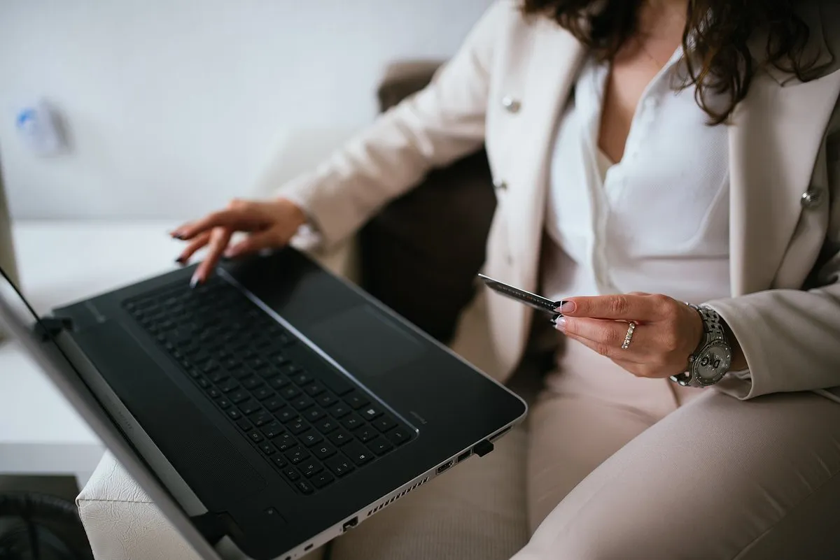 Kobieta w jasnym garniturze trzyma kartę płatniczą i wpisuje dane na laptopie, który ma na kolanach