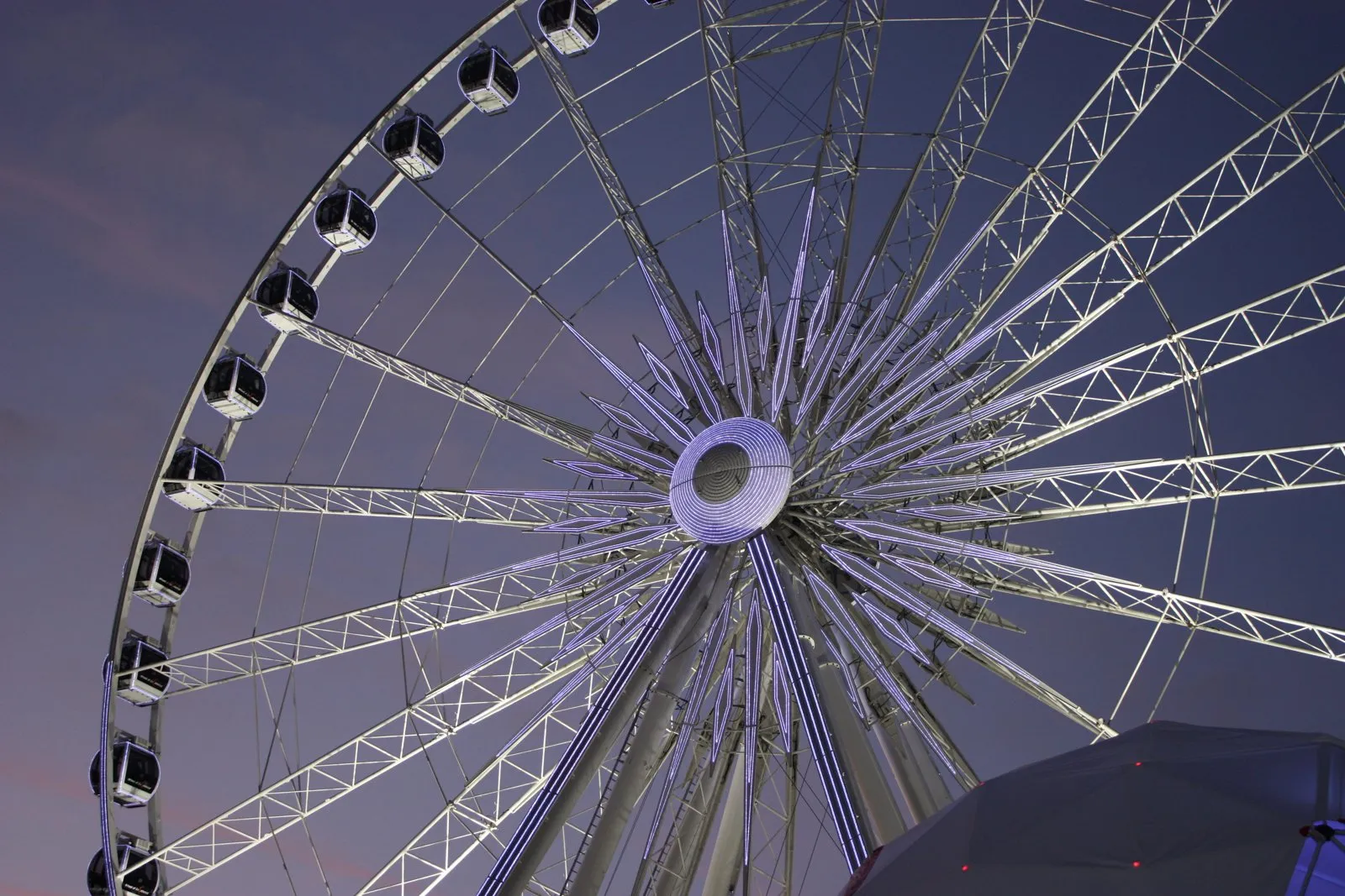Close-up of the steel structure of a Ferris wheel with its gondolas, the hub lit up in purple, at dusk