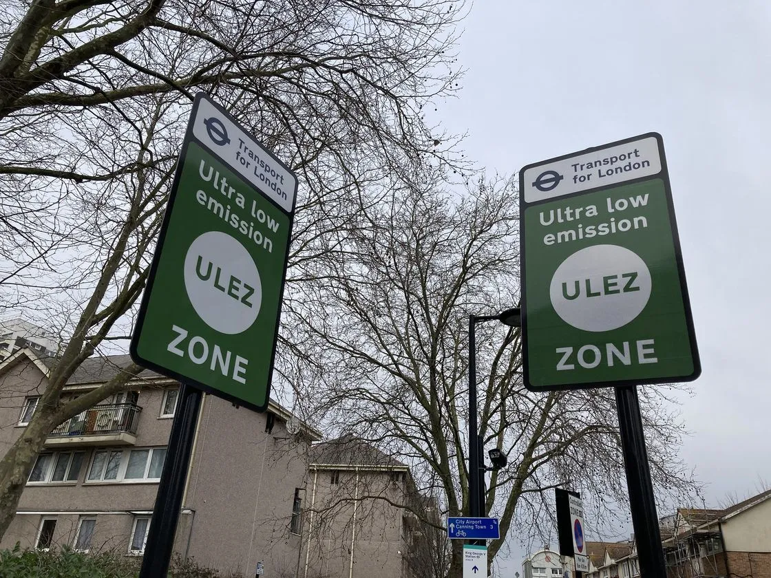Two green Transport for London signs reading Ultra low emission ULEZ zone against bare trees and a block of flats