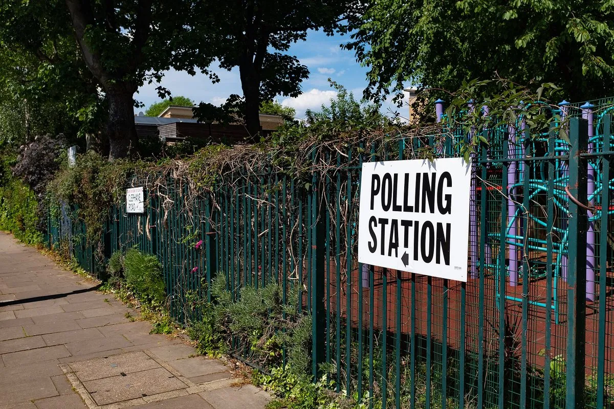 Biały znak z napisem Polling Station i strzałką, przymocowany do zielonego ogrodzenia przy londyńskim chodniku
