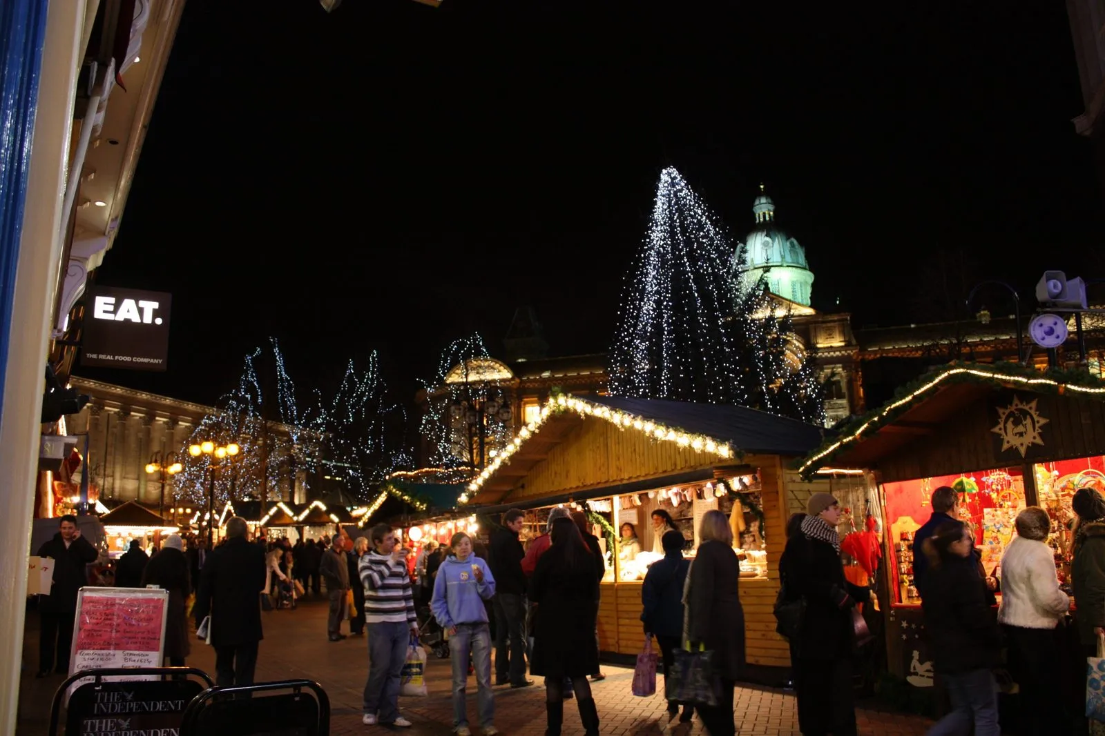 Jarmark Frankfurt Christmas Market w Victoria Square w Birmingham wieczorem: drewniane stragany z girlandami światełek, tłum kupujących, w tle oświetlona kopuła Council House i drzewa z lampkami