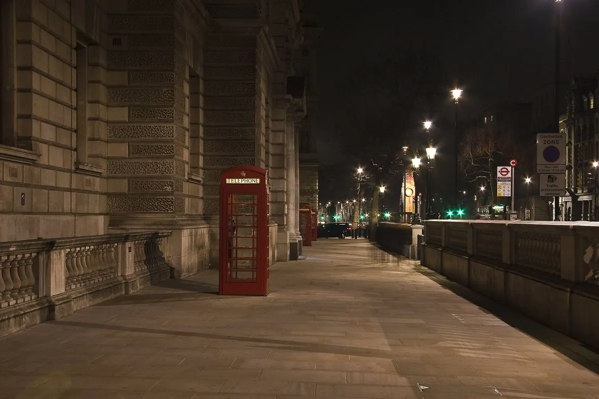 A red telephone box on an empty pavement next to a stone building in London at night