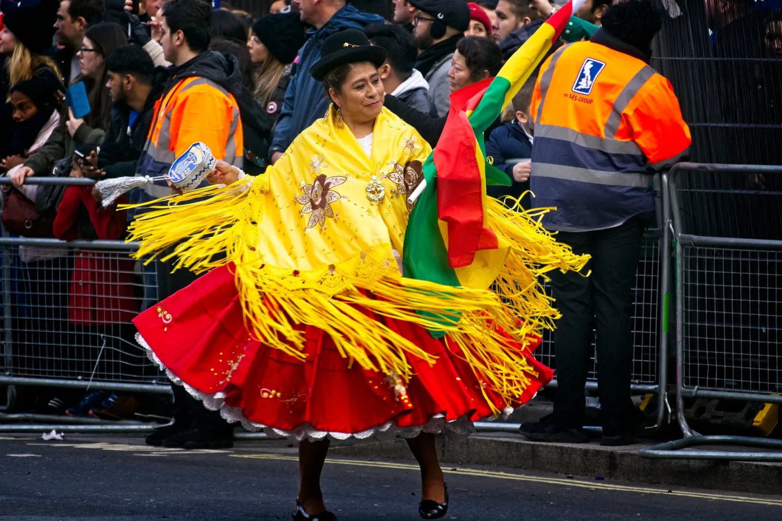 A dancer in a red skirt and a yellow fringed shawl, holding a Bolivian flag, dances in the street; behind the barriers there is a crowd of spectators and stewards in orange high-visibility vests
